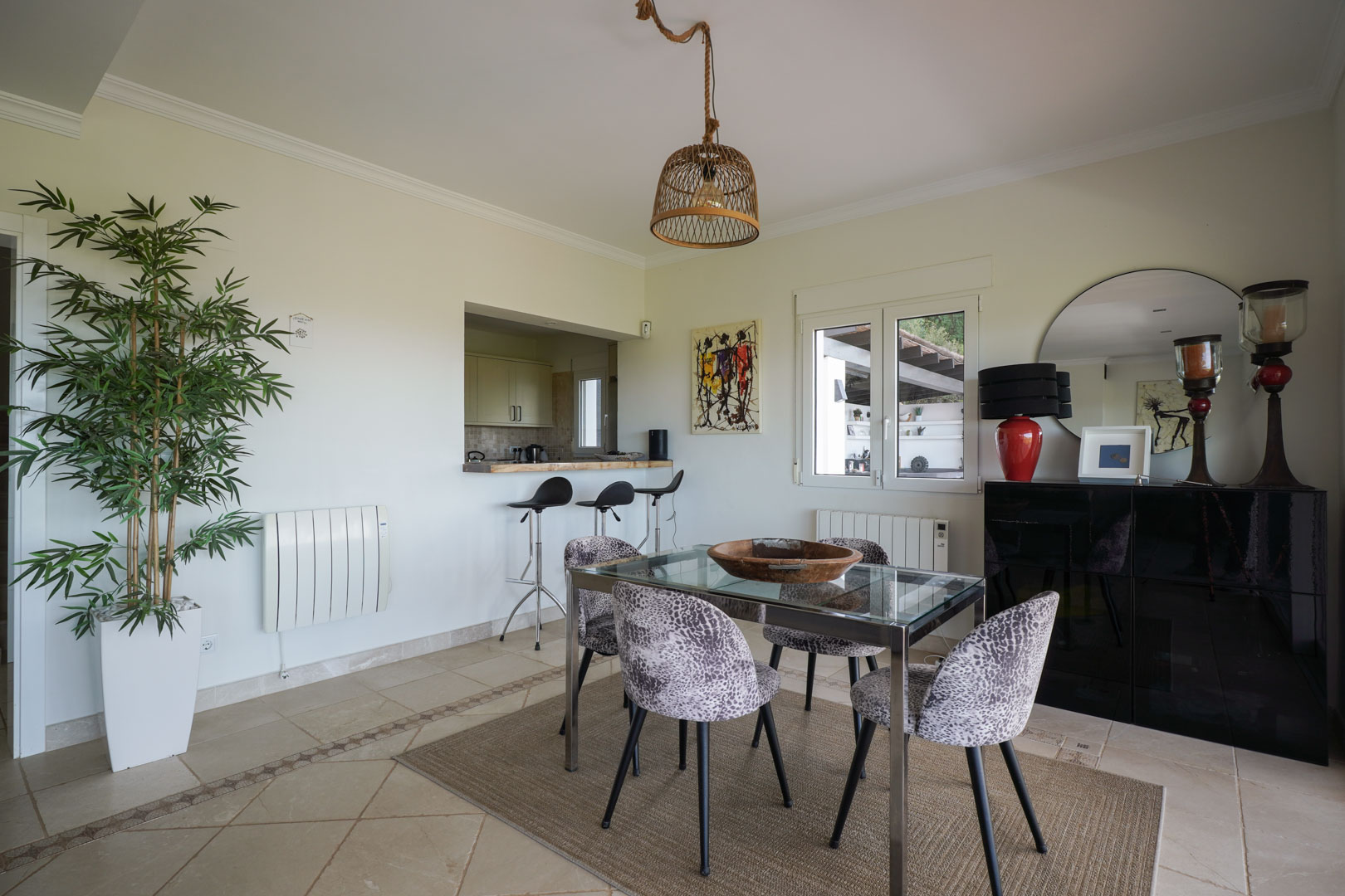 Lichte eethoek met glazen tafel, beklede stoelen en een rieten hanglamp, verbonden met de halfopen keuken via een ontbijtbar met krukken.
