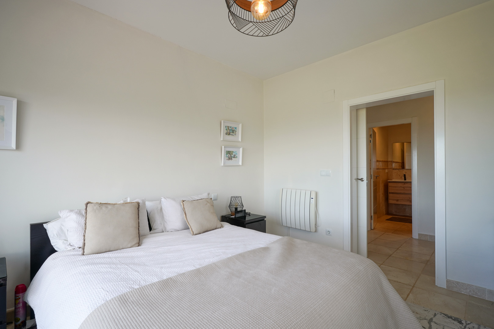 Lichte slaapkamer met keramische tegelvloer, tweepersoonsbed en directe toegang tot de en-suite badkamer. Voorzien van elektrische radiator en moderne draadlamp.