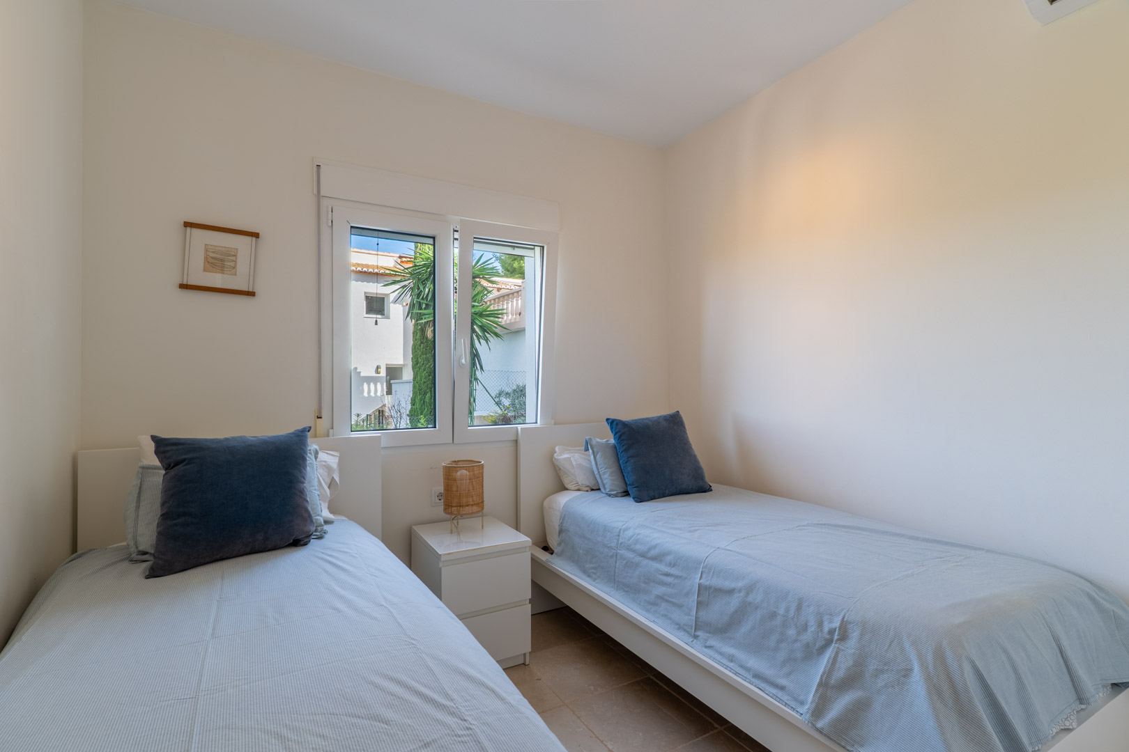 Slaapkamer met twee eenpersoonsbedden, wit meubilair en een raam dat zorgt voor natuurlijk licht. Voorzien van een praktische nachtkast en tegelvloer.