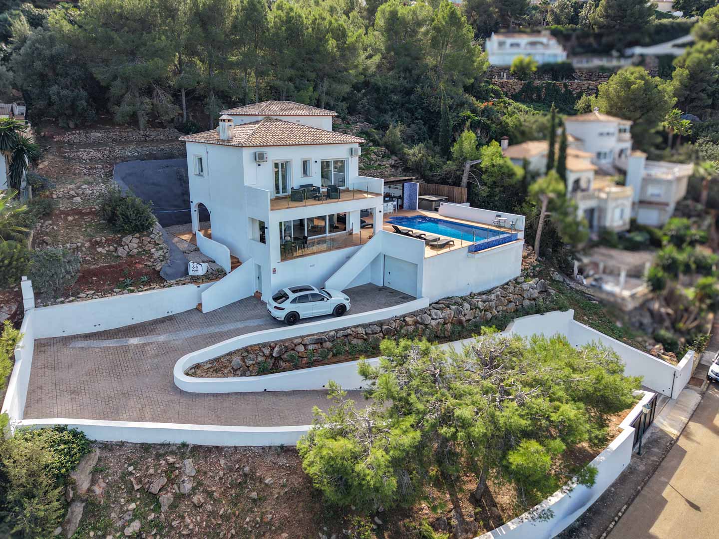 Verhoogd zicht op de villa met witte gevel, privézwembad, terrassen met glazen balustrades en een geplaveide oprit in de bosrijke heuvels van La Sella.