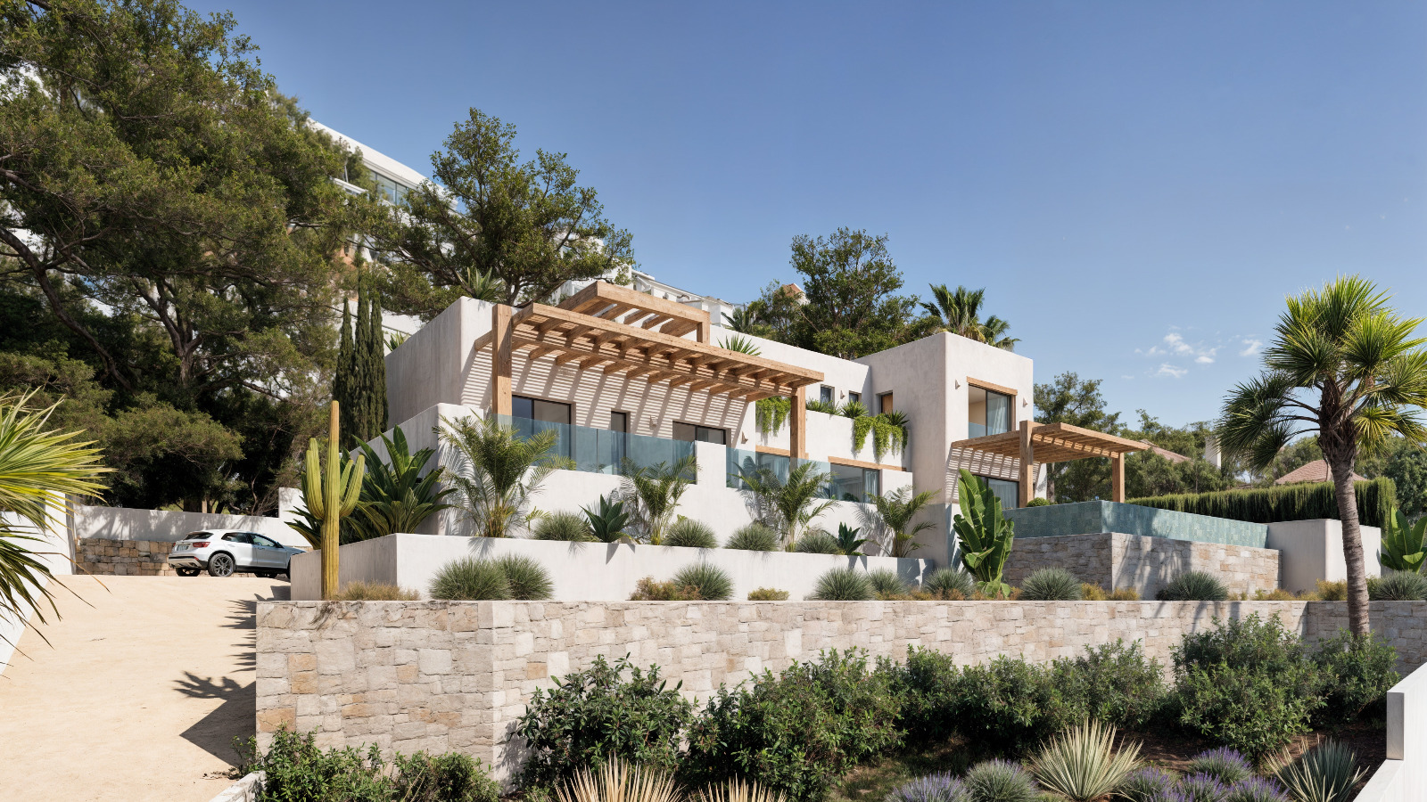 Buitenaanzicht van de villa in Ibizastijl met witte gevels, houten pergola's en natuurstenen muren geïntegreerd in een mediterrane tuin met palmbomen.