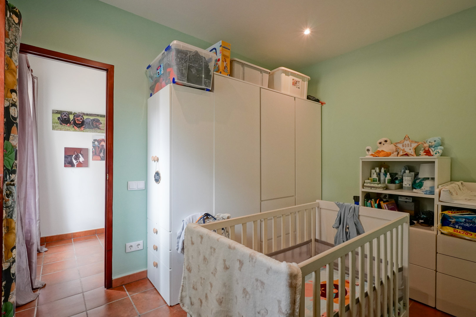 Babykamer met een wit houten wiegje, bijpassende kastenwand en lichtgroene muren. De ruimte is voorzien van een traditionele tegelvloer en inbouwspots in het plafond.