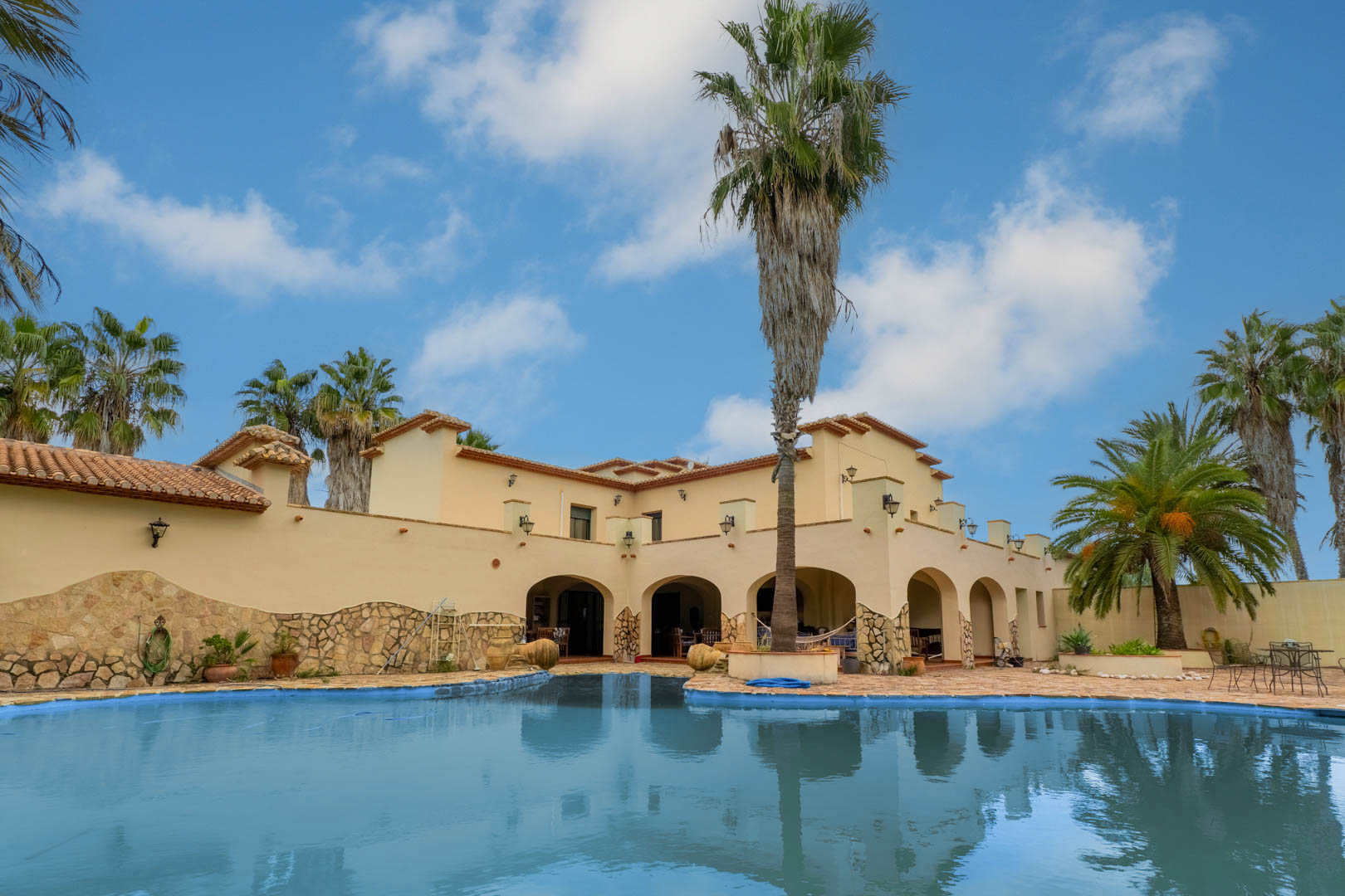Buitenaanzicht van het niervormige zwembad en het stenen terras, met de mediterrane gevel van de villa, boogvormige veranda's en hoge palmbomen.
