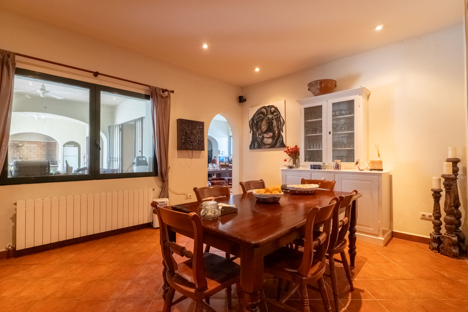 Eethoek met terracotta vloertegels, een massief houten tafel en een witte buffetkast. Het grote raam biedt uitzicht op de veranda naast de boogdoorgang.