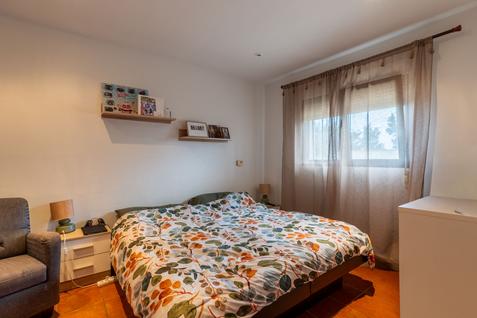 Lichte slaapkamer met terracotta tegelvloer, witte muren en houten wandplanken. Een raam met lichtdoorlatende gordijnen en een rolluik zorgt voor natuurlijk daglicht.