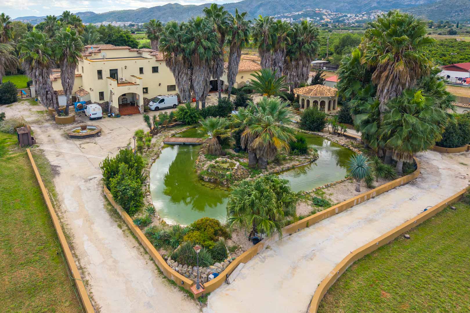 Luchtfoto van de hoofdingang met een grote siervijver, een houten brug en volwassen palmbomen voor de mediterrane villa.