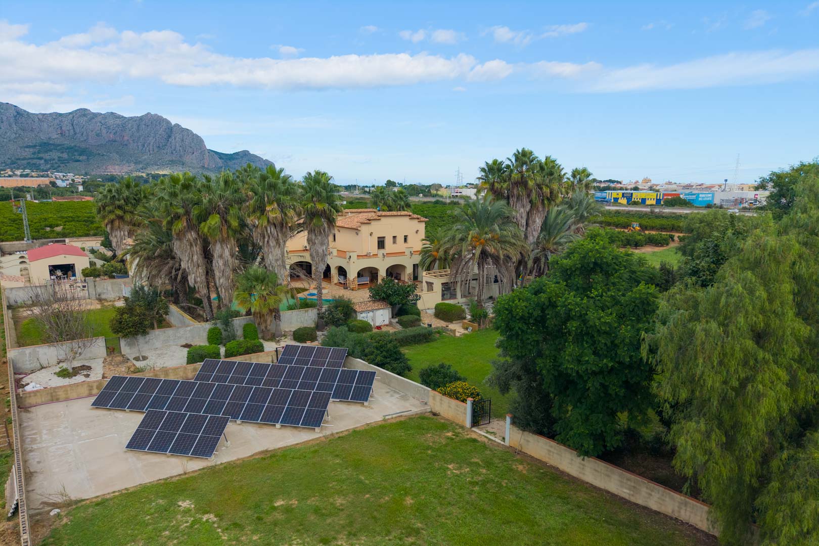 Luchtfoto van het landgoed met zonnepanelen, uitgestrekte gazons, volgroeide palmbomen en de mediterrane villa tegen een bergachtig decor.