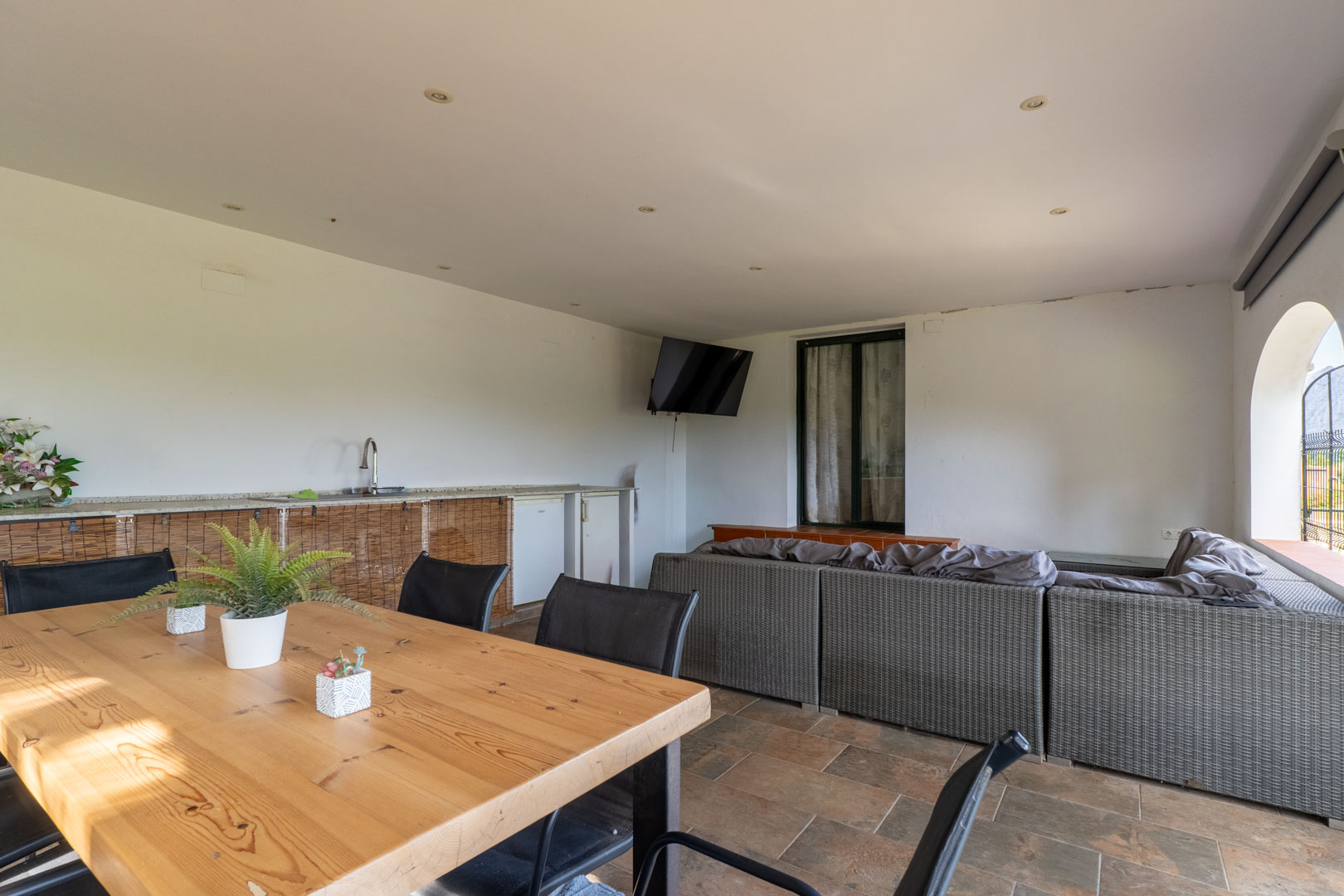 Overdekt terras met een grijze rotan zithoek, een massief houten eettafel en een buitenkeuken. De ruimte is afgewerkt met inbouwspots en een rustieke tegelvloer.