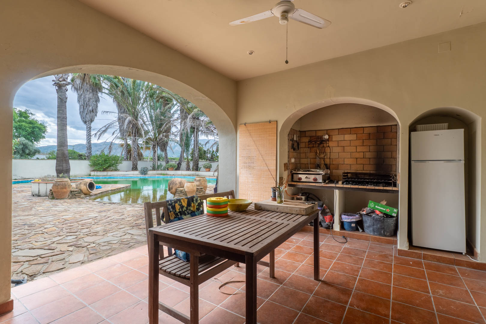 Overdekt terras met een ingebouwde stenen barbecue, houten eethoek en uitzicht op het zwembad en de tuinen via een karakteristieke boog.