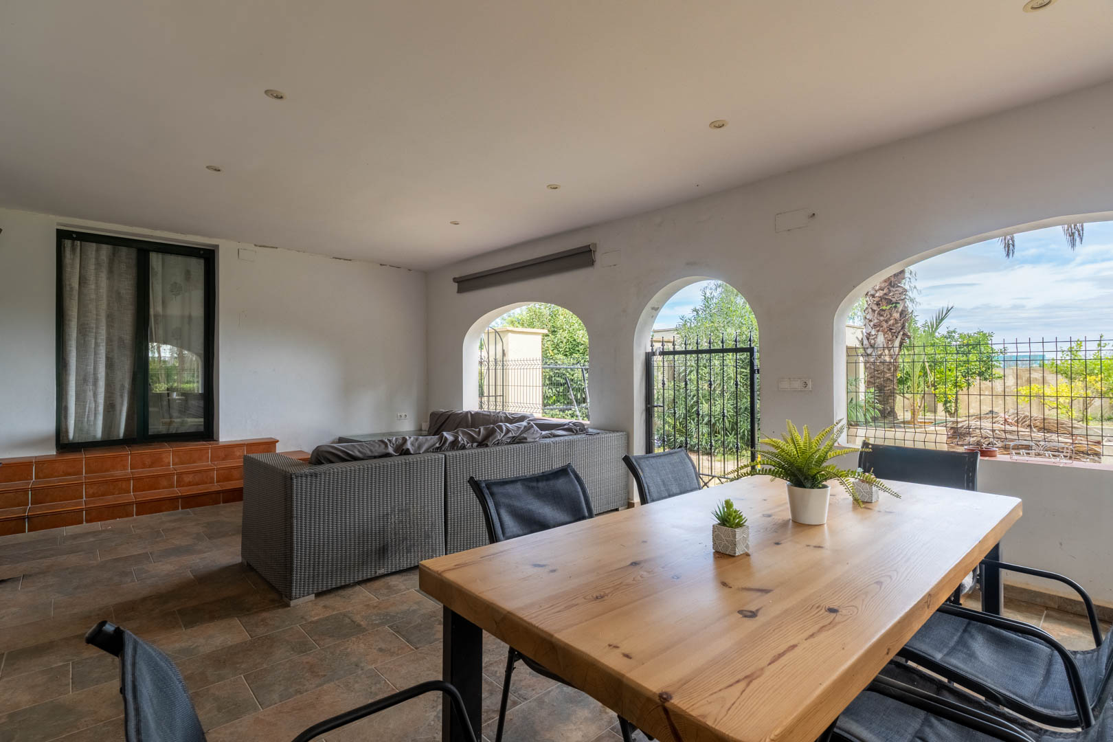 Overdekt terras met mediterrane bogen, massief houten eettafel, rotan banken en uitzicht op de tuin. Rustieke tegelvloer en inbouwspots in het plafond.