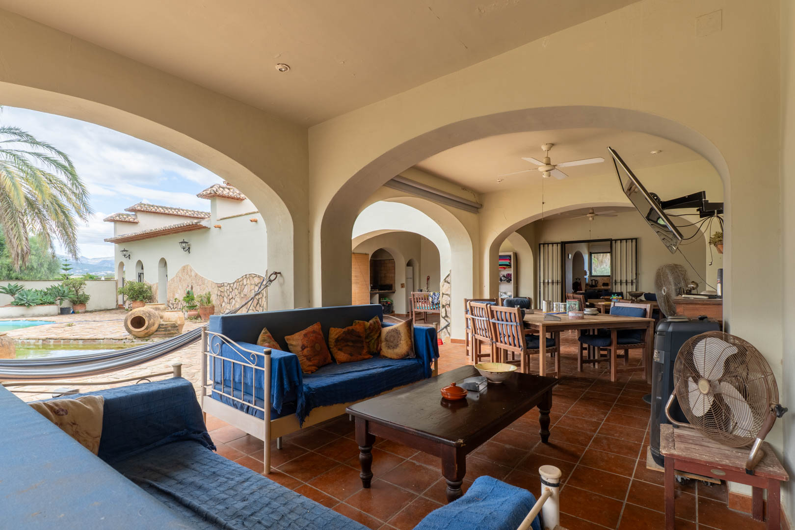 Overdekt terras met mediterrane bogen, terracotta vloer en een zithoek. Het biedt uitzicht op het zwembad, de hangmat en de omliggende tuinen met palmbomen.