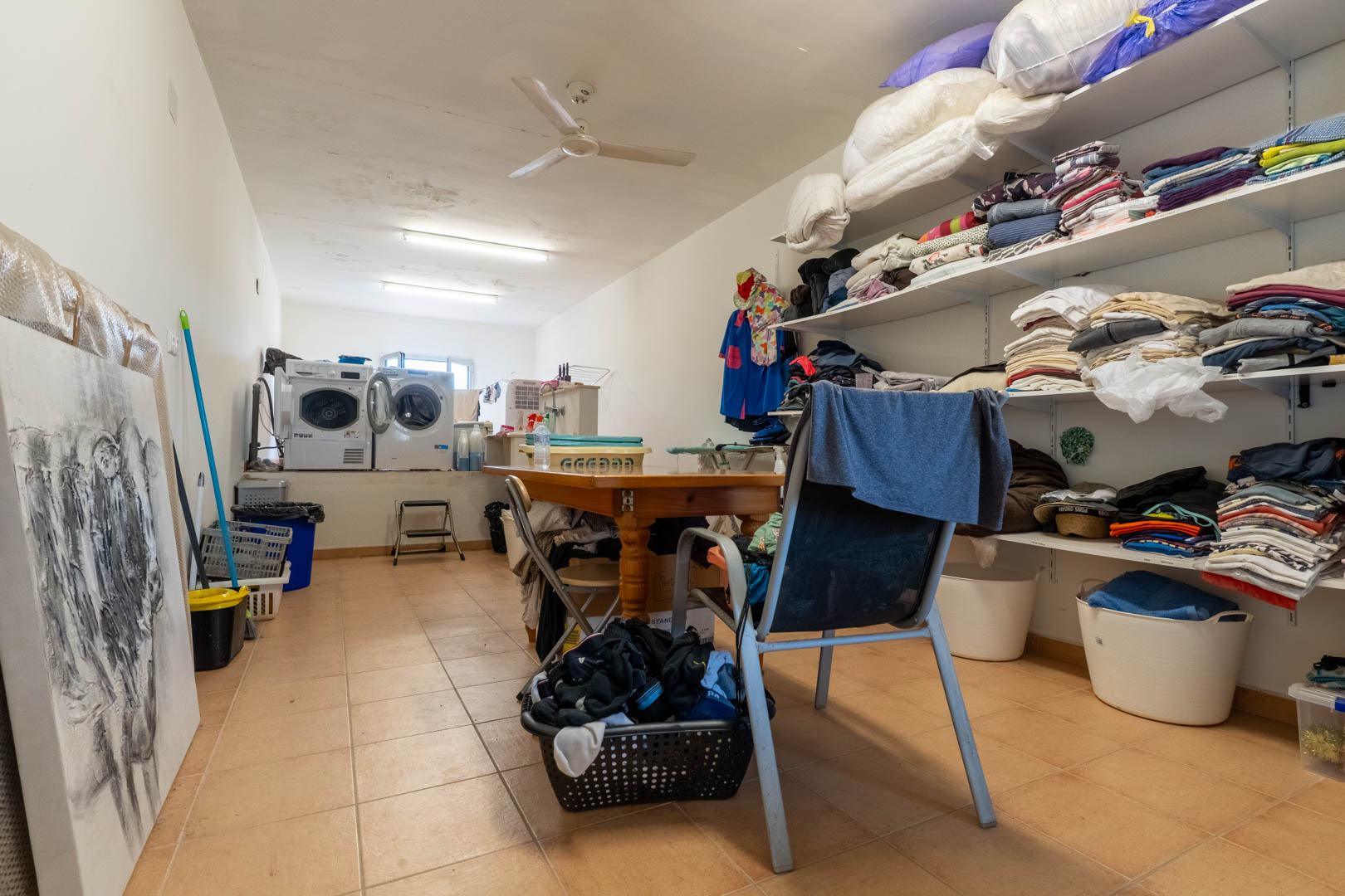 Ruime wasruimte en berging met verhoogde wasmachines, uitgebreide open stellingen voor linnengoed en een centrale houten werktafel.