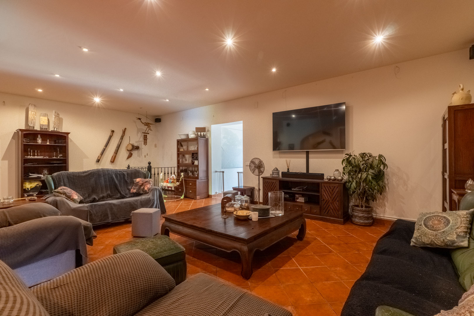 Ruime woonkamer met rustieke terracotta vloertegels, inbouwspots in het plafond en massief houten meubilair. De ruimte beschikt over een mediahoek en toegang tot de trap.