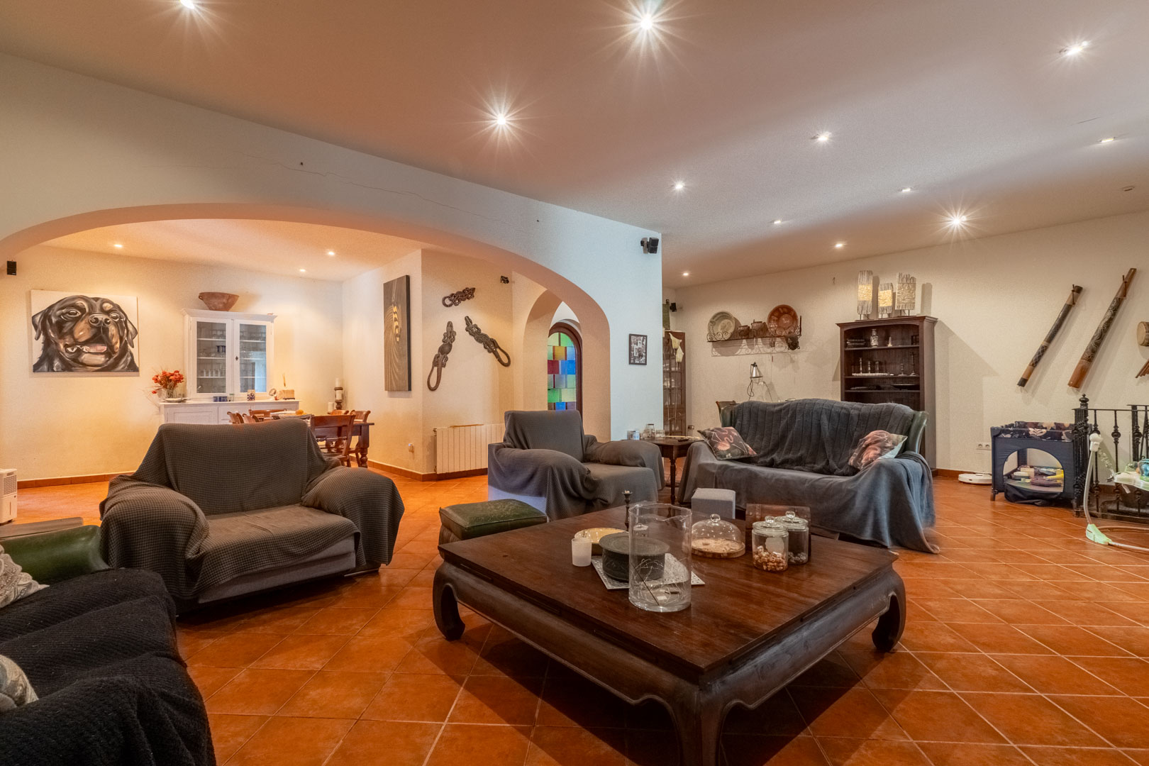 Ruime woonkamer met terracotta tegelvloer, inbouwspots in het plafond en een grote boogverbinding naar de aangrenzende eetkamer en hal.