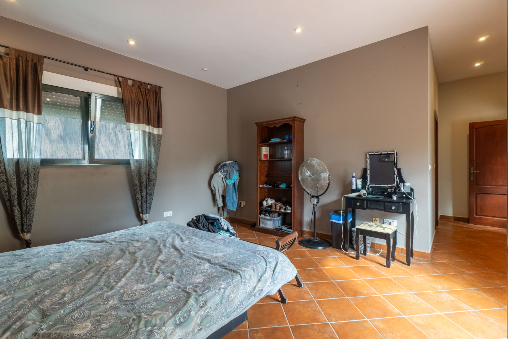Slaapkamer met diagonaal gelegde terracotta tegelvloer, inbouwspots en muren in aardetinten, voorzien van een raam met rolluik.