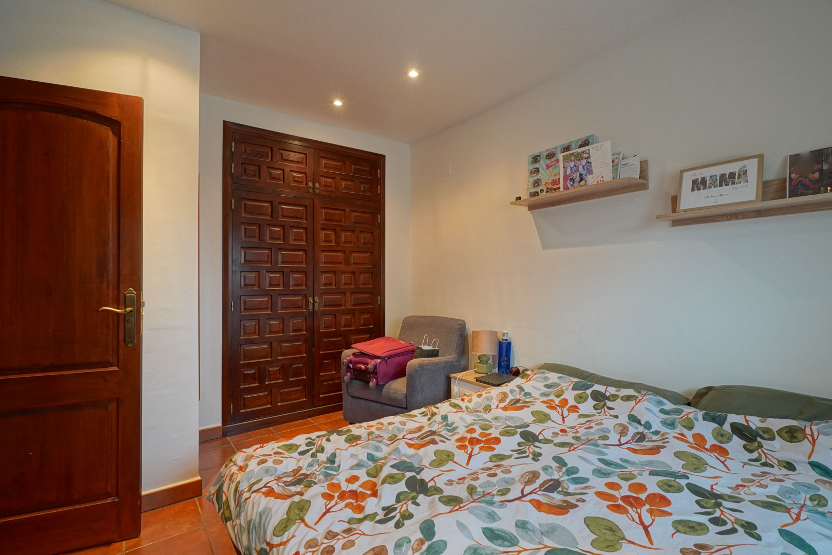 Slaapkamer met een ingebouwde houten kledingkast in traditionele stijl, terracotta vloertegels en subtiele inbouwspots in het plafond.