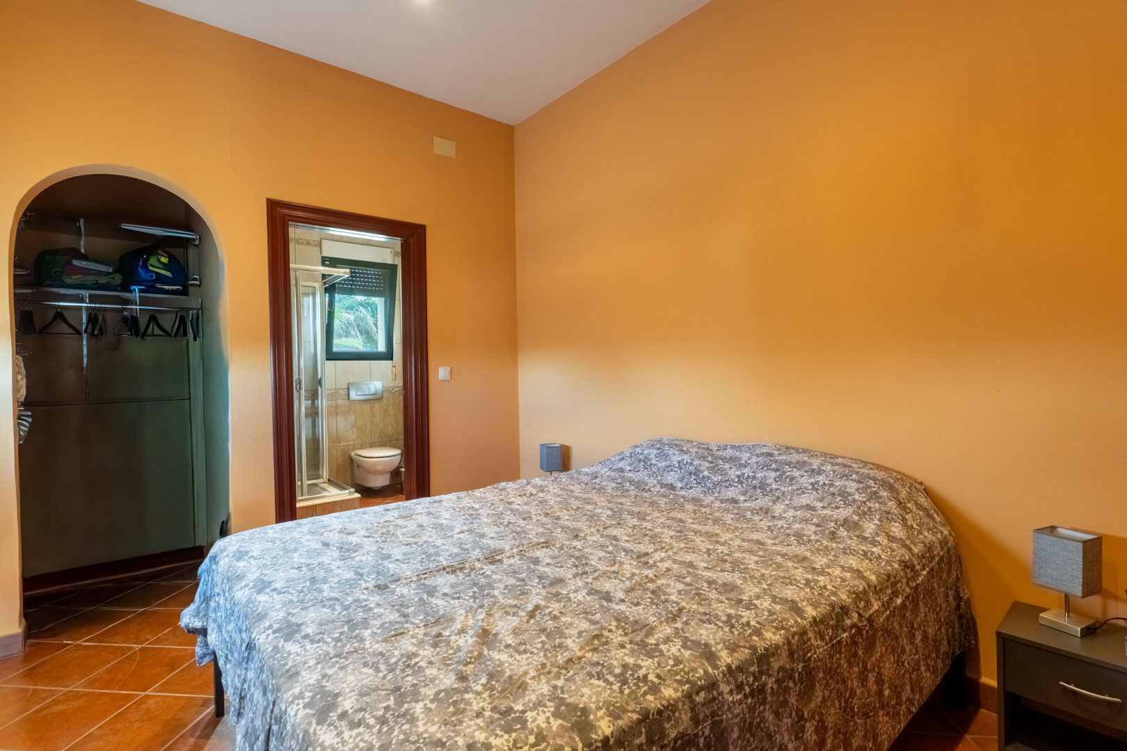 Slaapkamer met terracotta vloer en okergele muren, voorzien van een inloopkast met boog en een aangrenzende badkamer met douche.