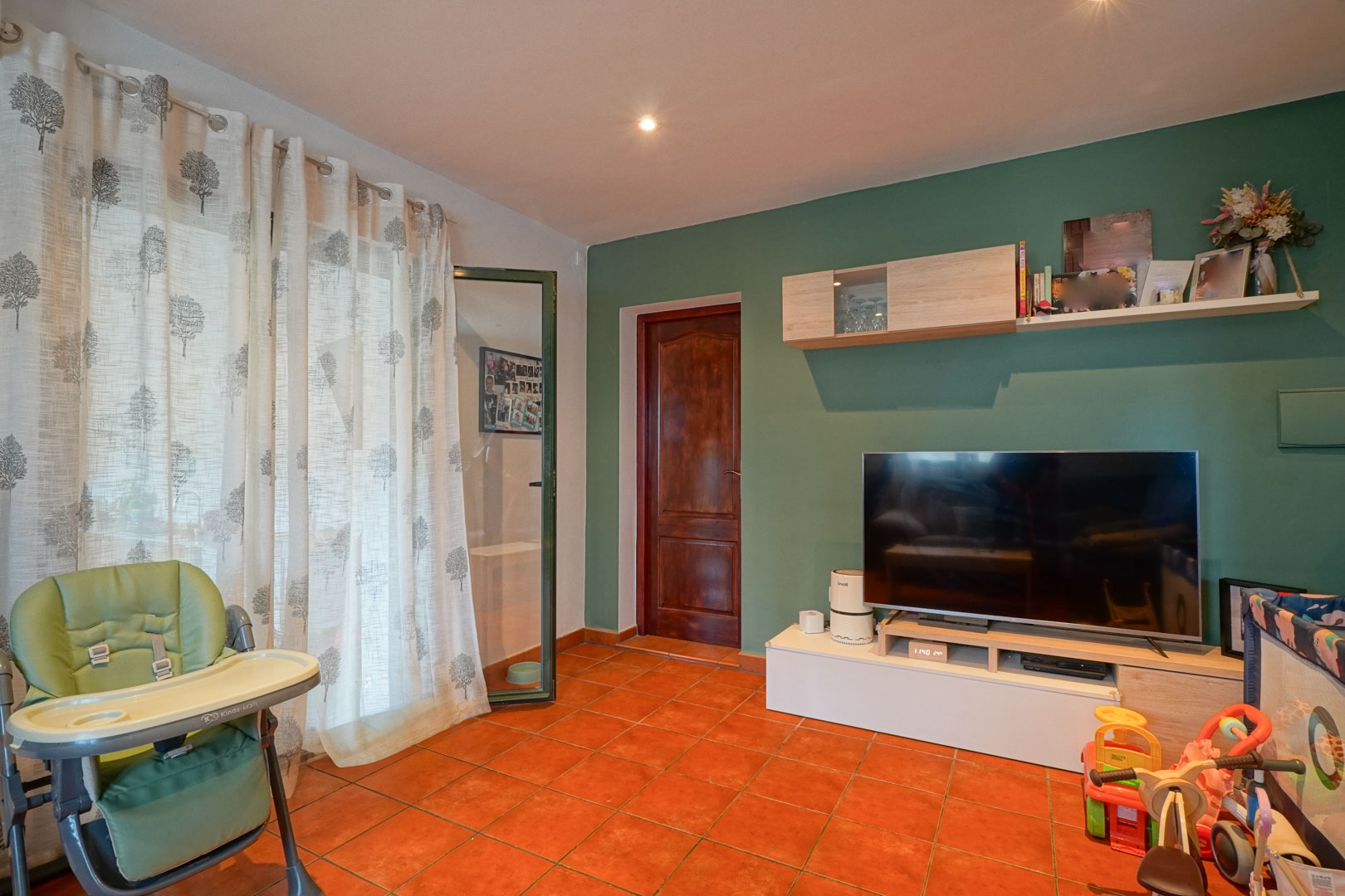 Woonkamer met een saliegroene accentmuur, terracotta tegelvloer en een modern tv-meubel. Een houten binnendeur en inbouwspots zorgen voor een warme afwerking.