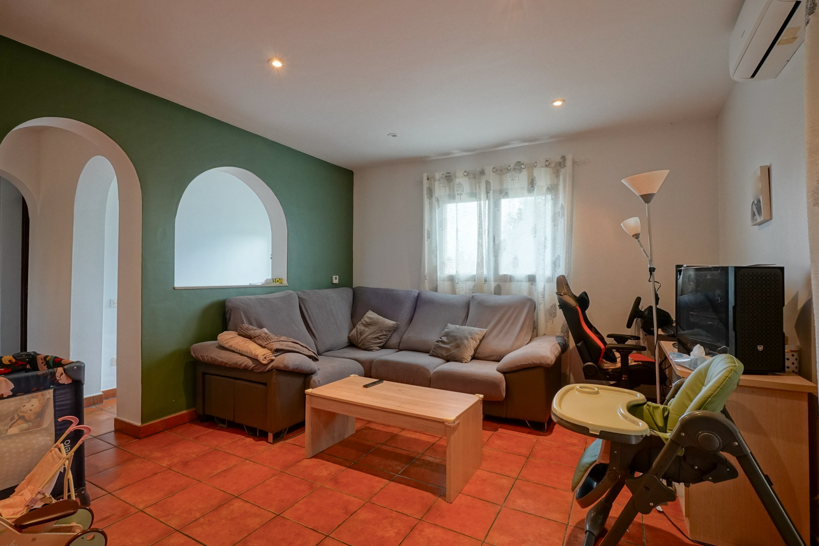 Woonkamer met terracotta vloertegels, groene accentmuur en ronde boogopeningen. Voorzien van een hoekbank, bureauhoek en split-airconditioning.