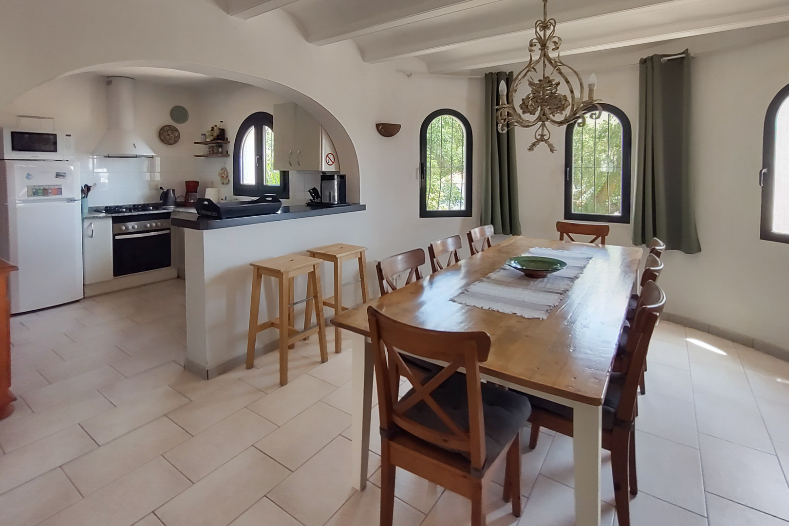 Eethoek met een grote houten tafel voor acht personen, verbonden via een boog met een open keuken voorzien van een ontbijtbar, krukken en halfronde ramen.