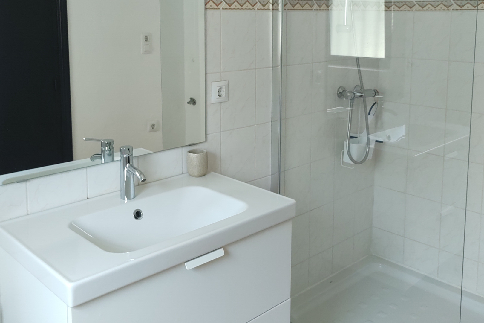 Functionele badkamer met wit wastafelmeubel, chromen kranen en een inloopdouche met glazen wand. Witte betegelde wanden met een decoratieve sierrand bovenaan.