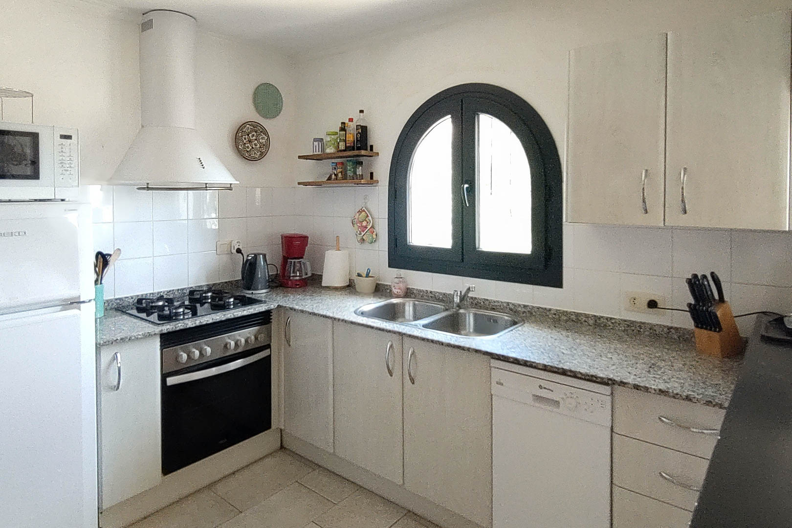 Functionele keuken met granieten werkblad, gasfornuis en witte kasten. Een karakteristiek boograam met donker kozijn biedt natuurlijk licht boven de dubbele spoelbak.