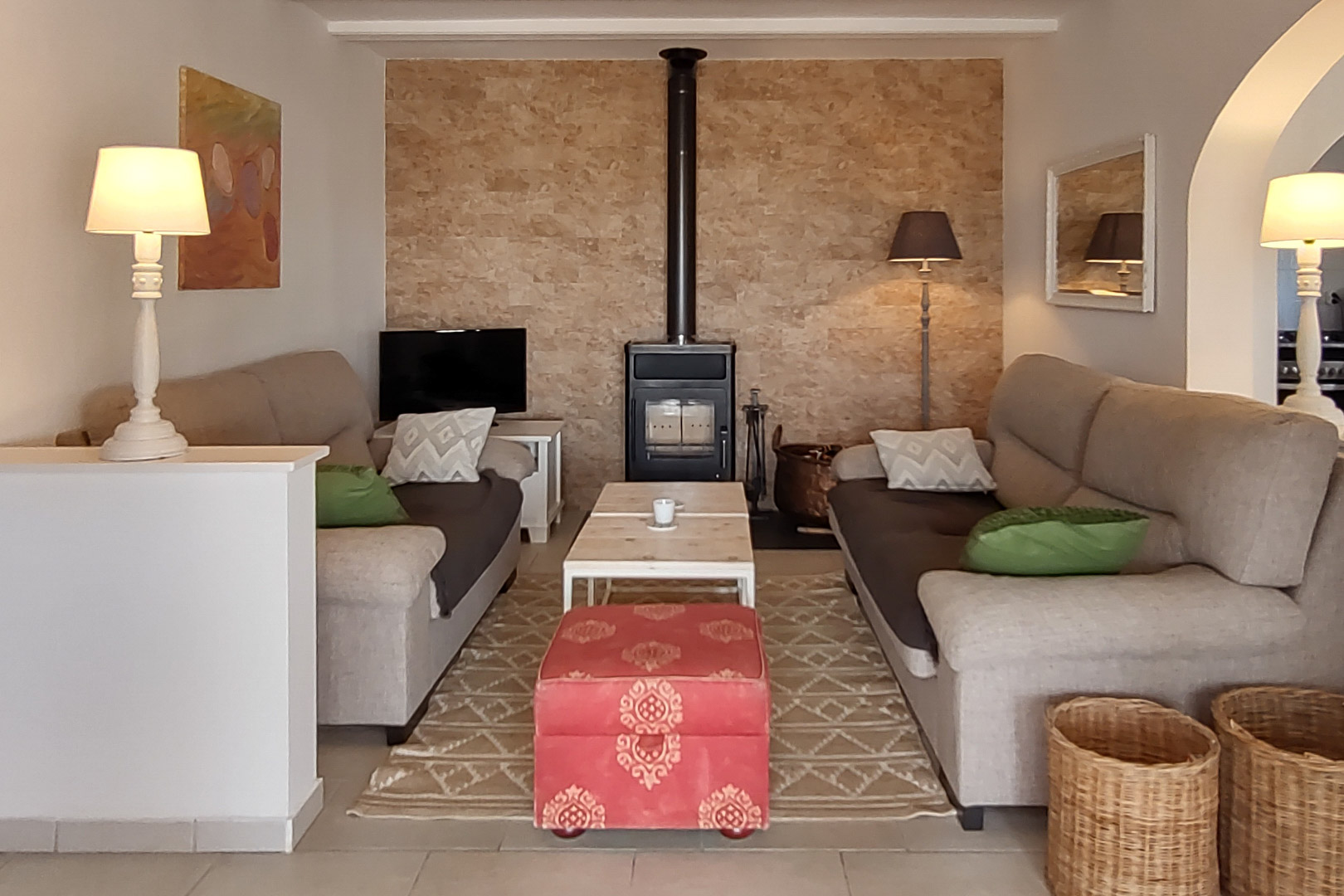 Gezellige woonkamer met houtkachel tegen een natuurstenen wand, tegenover elkaar geplaatste banken, houten salontafel en een rode decoratieve hocker.
