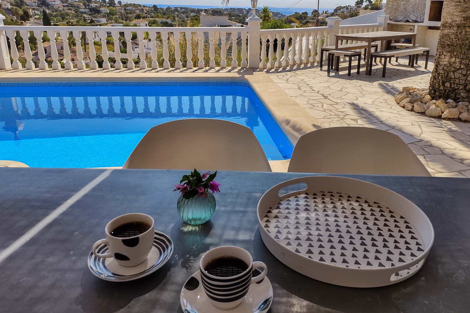 Hoofdterras met privézwembad en uitzicht op zee. Inclusief een eethoek buiten met houten tafel en banken op natuurstenen bestrating en een witte balustrade.