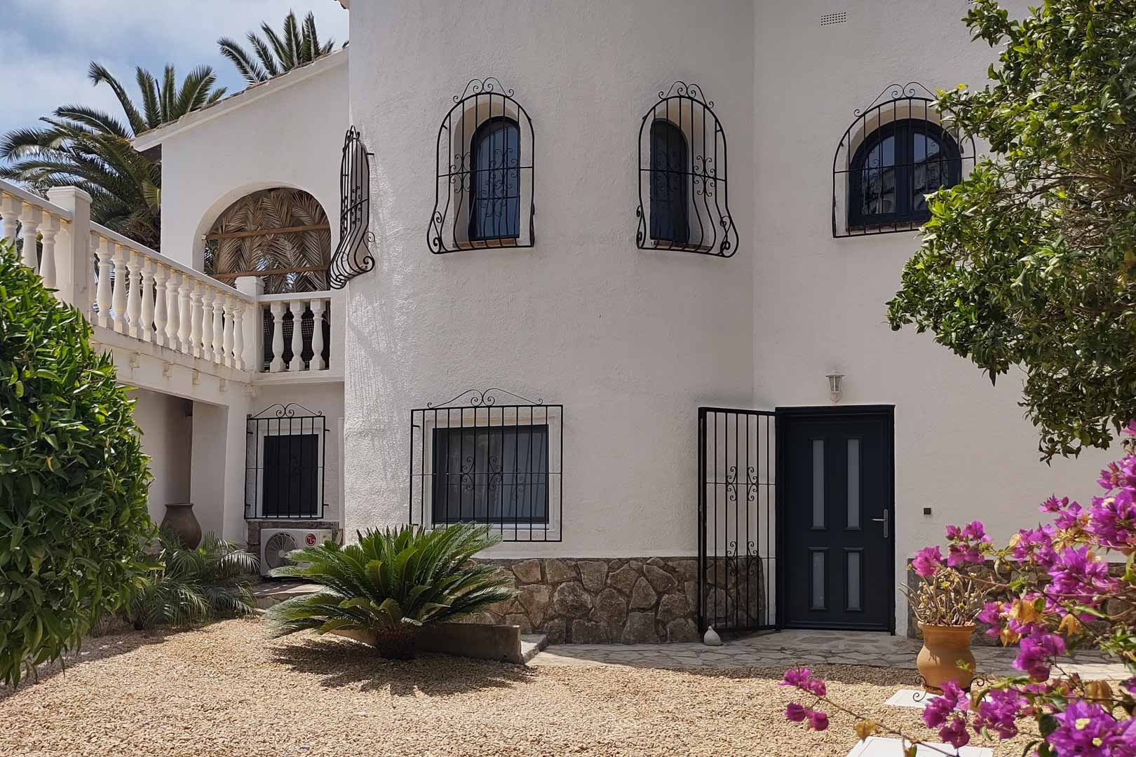 Mediterrane witte gevel met stenen plint, ramen voorzien van smeedijzeren traliewerk en een hoofdingang grenzend aan een grindtuin met kleurrijke bougainville.