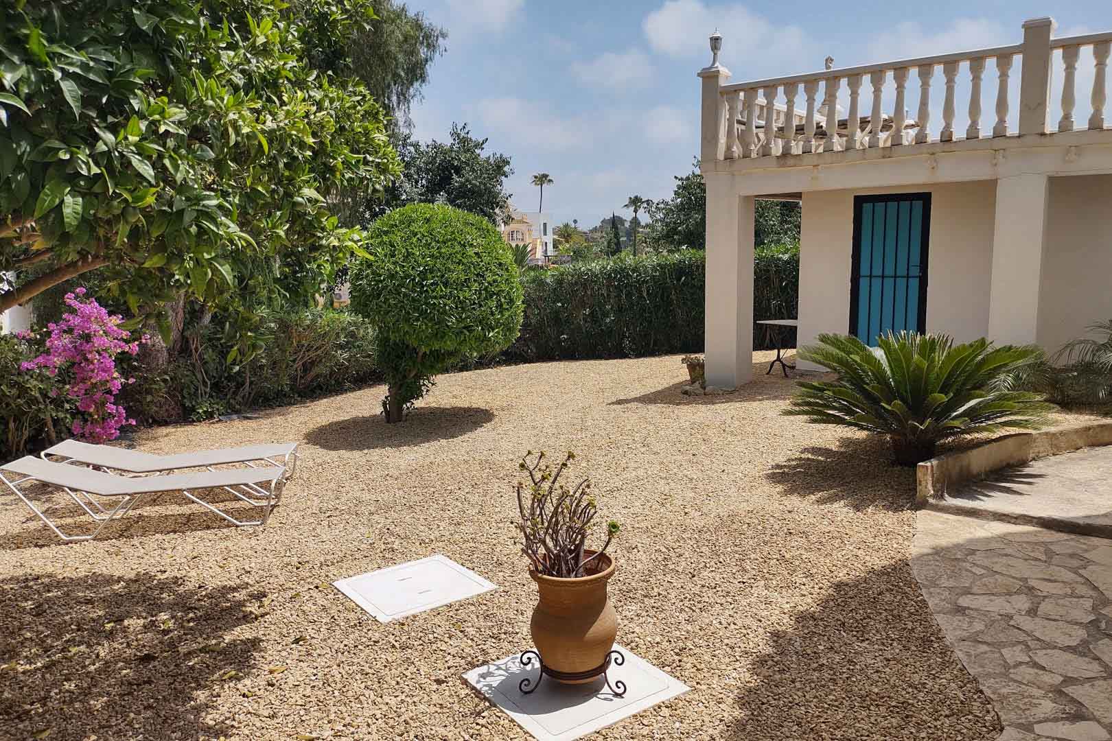Onderhoudsvriendelijke mediterrane tuin met grind, ligbedden, lokale beplanting en een witte gevel met klassieke stenen balustrade en blauwe deur.