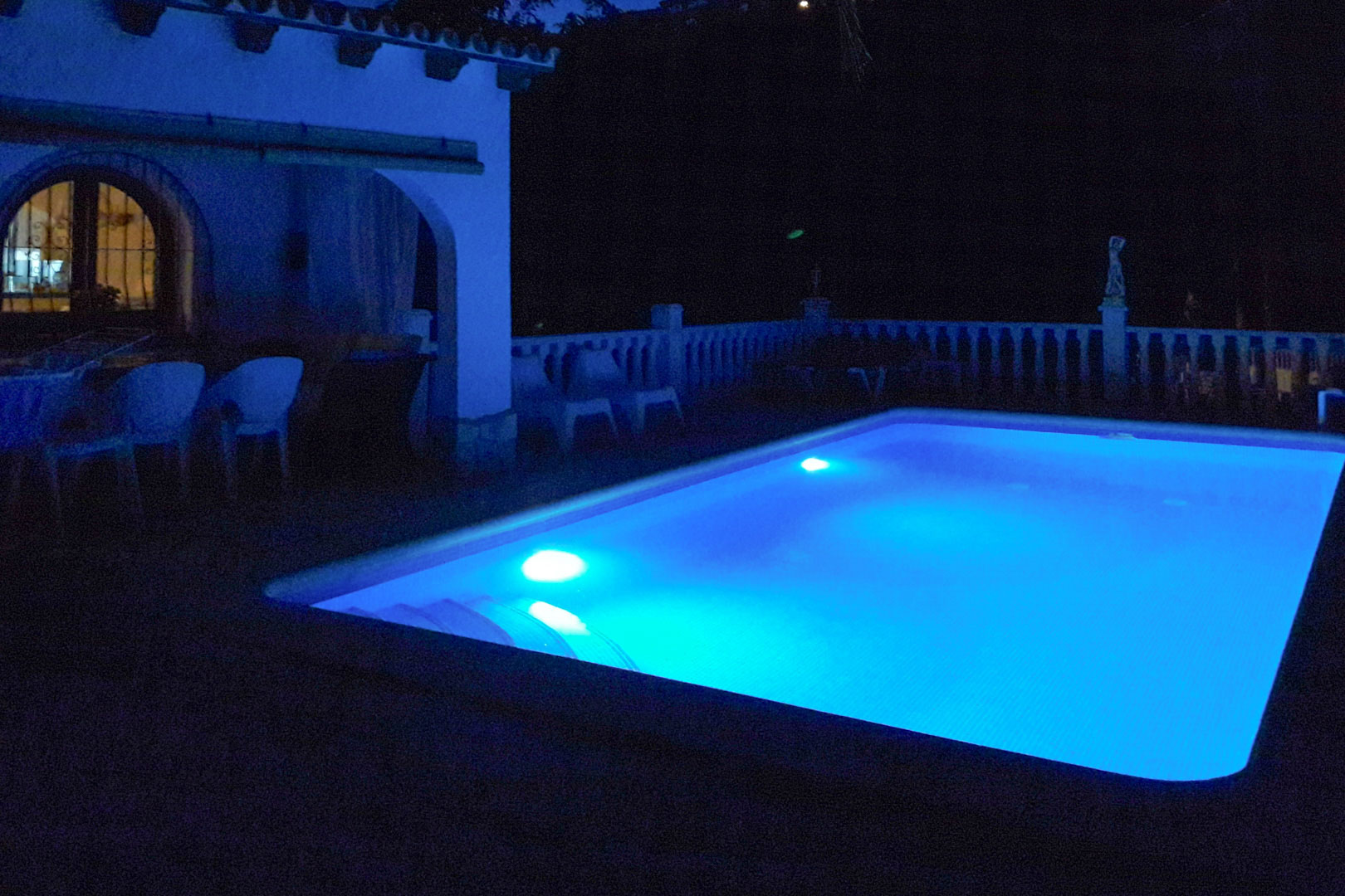 Rechthoekig zwembad met blauwe LED-onderwaterverlichting, gelegen naast een overdekt terras met bogen en een klassieke witte stenen balustrade.