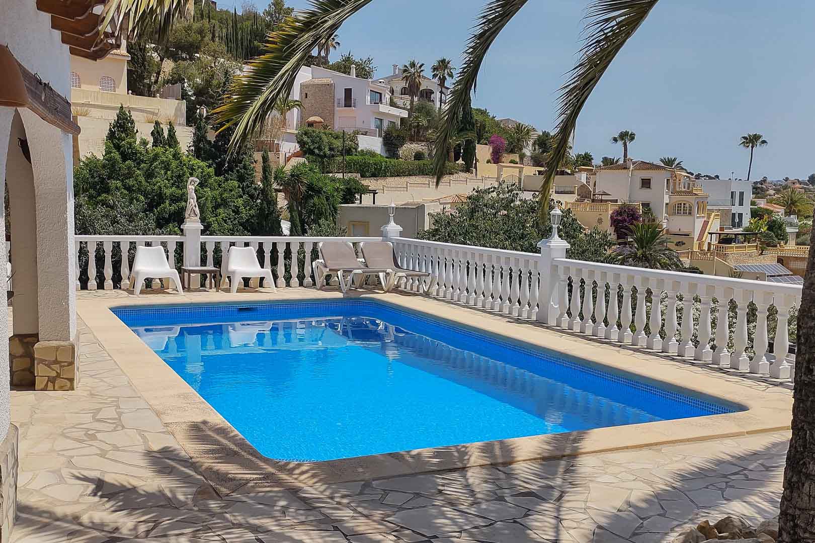 Rechthoekig zwembad met blauwe tegels, omgeven door een terras van natuursteen en een witte balustrade met uitzicht op de omliggende villa's en palmen.