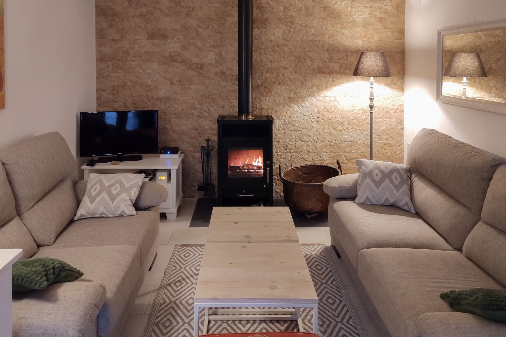 Sfeervolle woonkamer met houtkachel tegen een natuurstenen wand, ingericht met neutrale banken en een licht houten salontafel.