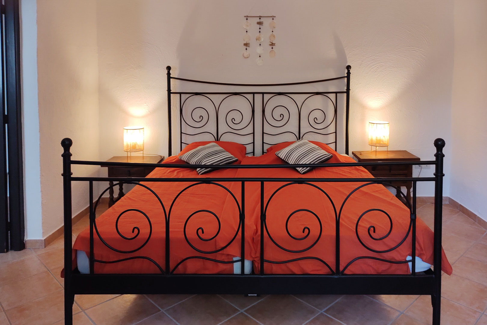 Slaapkamer met een zwart smeedijzeren bedframe, houten nachtkastjes en een keramische tegelvloer. Warme bedlampjes verlichten de witte muren met textuur.