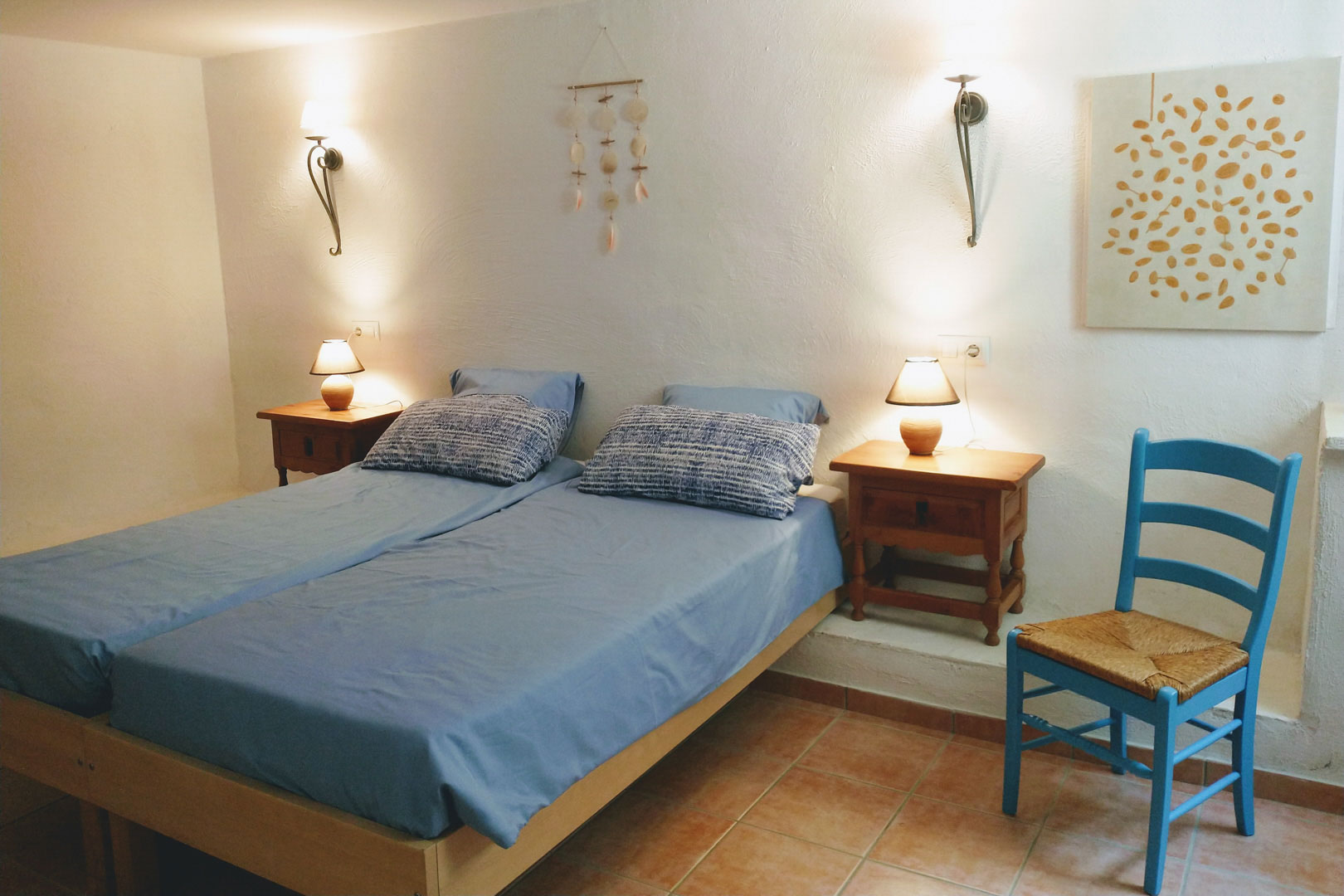 Slaapkamer met twee eenpersoonsbedden, terracotta vloertegels en witte muren. Voorzien van houten nachtkastjes, wandlampen en een blauwe rustieke stoel.