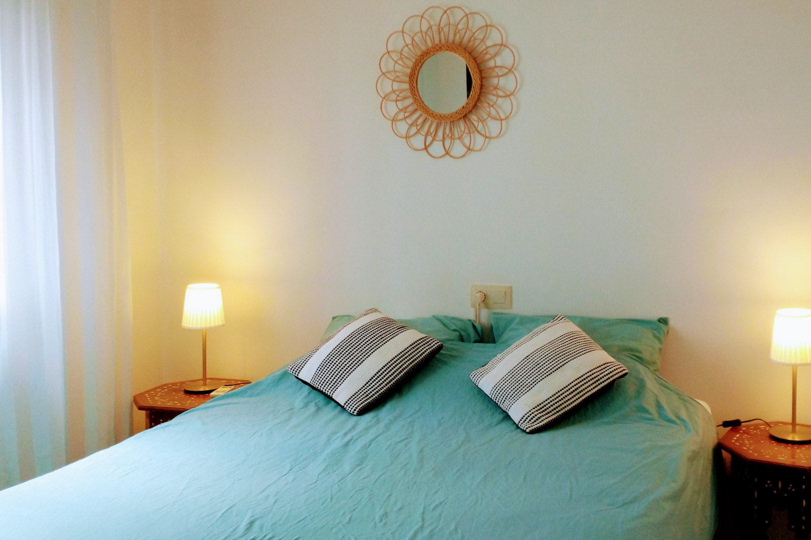 Slaapkamer met tweepersoonsbed, turquoise beddengoed, zeshoekige houten nachtkastjes en een decoratieve rotan spiegel aan de witte wand.