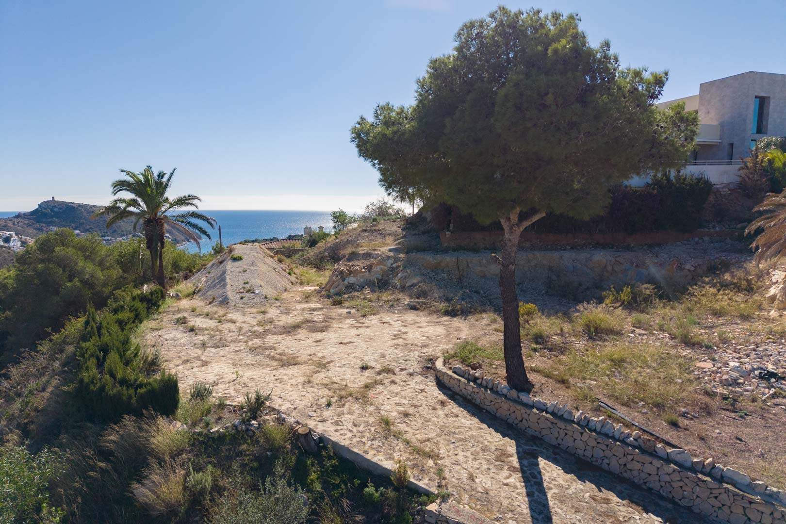 Buitenaanzicht van het perceel met natuurstenen pad, pijnbomen, palmen en panoramisch uitzicht op de Middellandse Zee in Moraira.