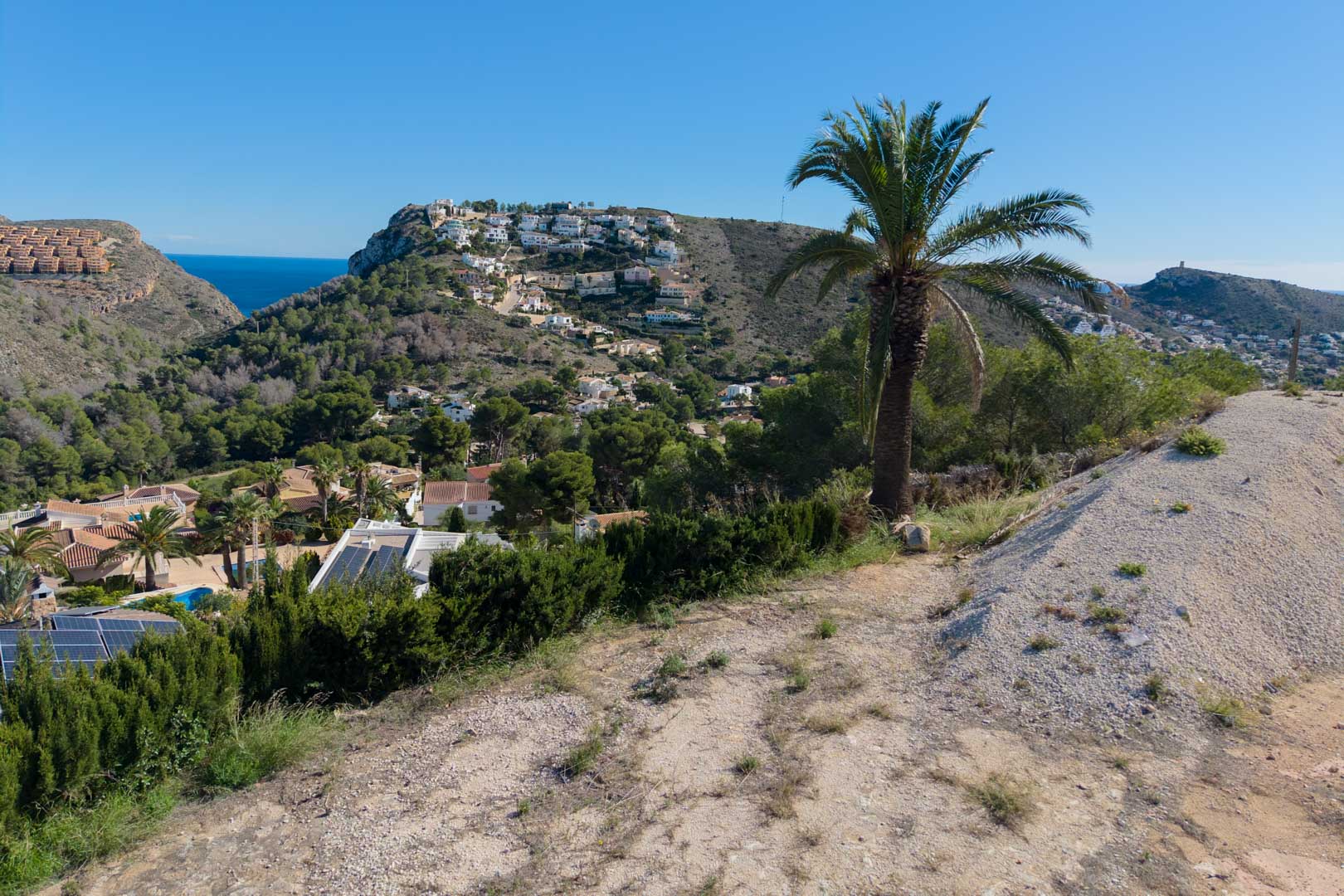 Panoramisch uitzicht vanaf het hoger gelegen perceel over de vallei van Moraira, met de Middellandse Zee op de achtergrond en heuvels vol dennen en palmen.