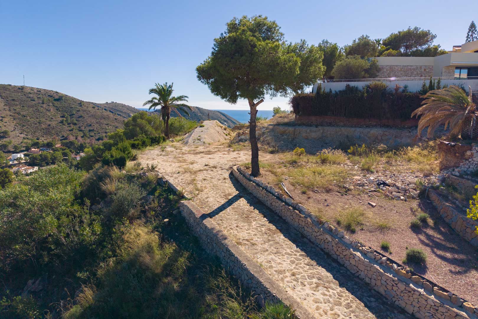 Panoramisch uitzicht vanaf het perceel met een geplaveid pad en droge stenen muren, omgeven door mediterrane flora en vrij uitzicht op zee.