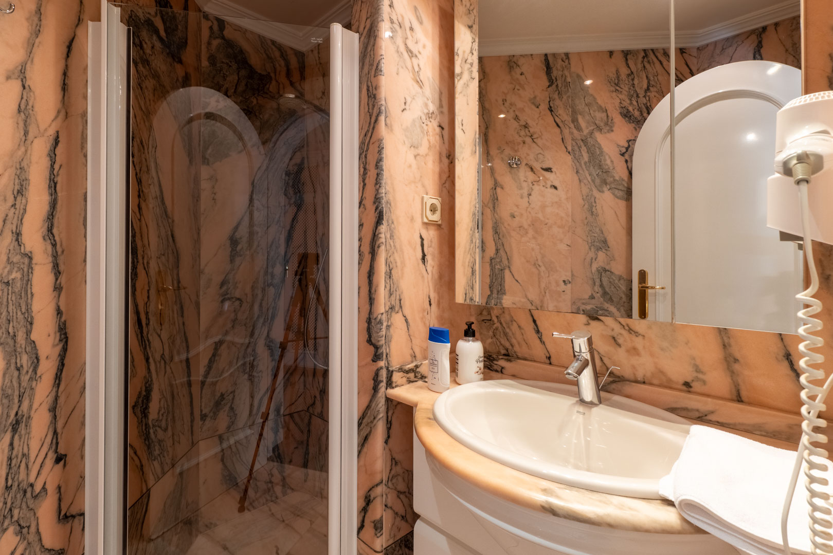 Badkamer met roze marmeren wanden en donkere aders, voorzien van een glazen douchecabine, een gebogen wastafelmeubel met natuurstenen blad en een grote spiegel.