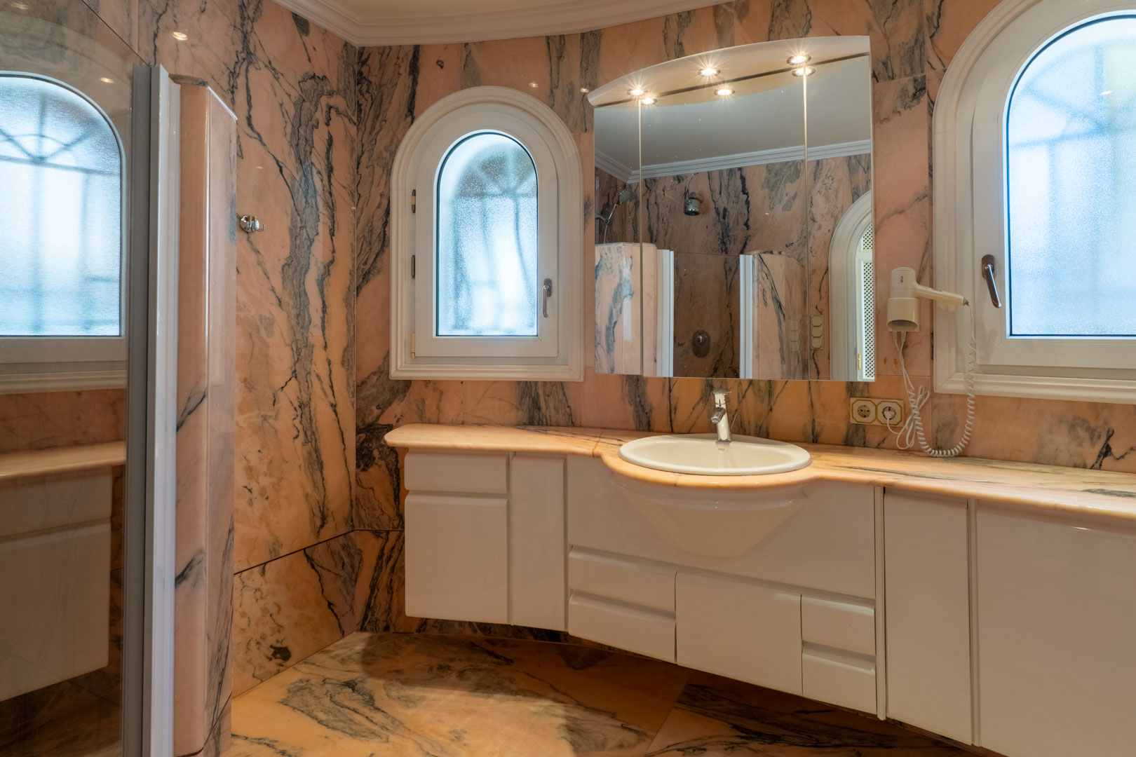 Badkamer met roze marmeren wanden en vloeren, een wit wastafelmeubel, een spiegel met spotjes en karakteristieke boogramen met matglas.