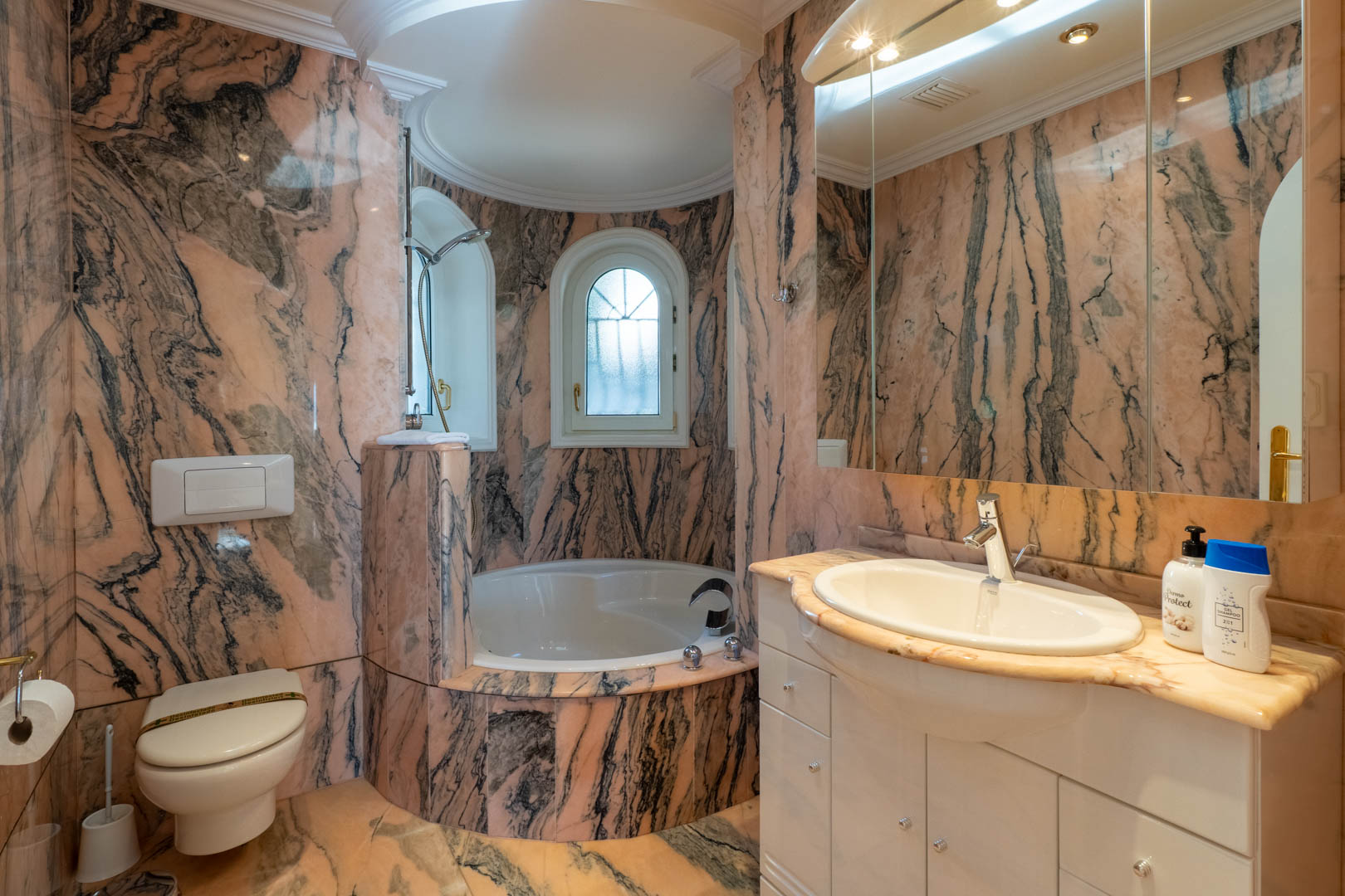 Badkamer met wanden en vloeren van roze en grijs geaderd marmer, voorzien van een rond ligbad in een nis, boogvensters en een wit wastafelmeubel met natuurstenen blad.