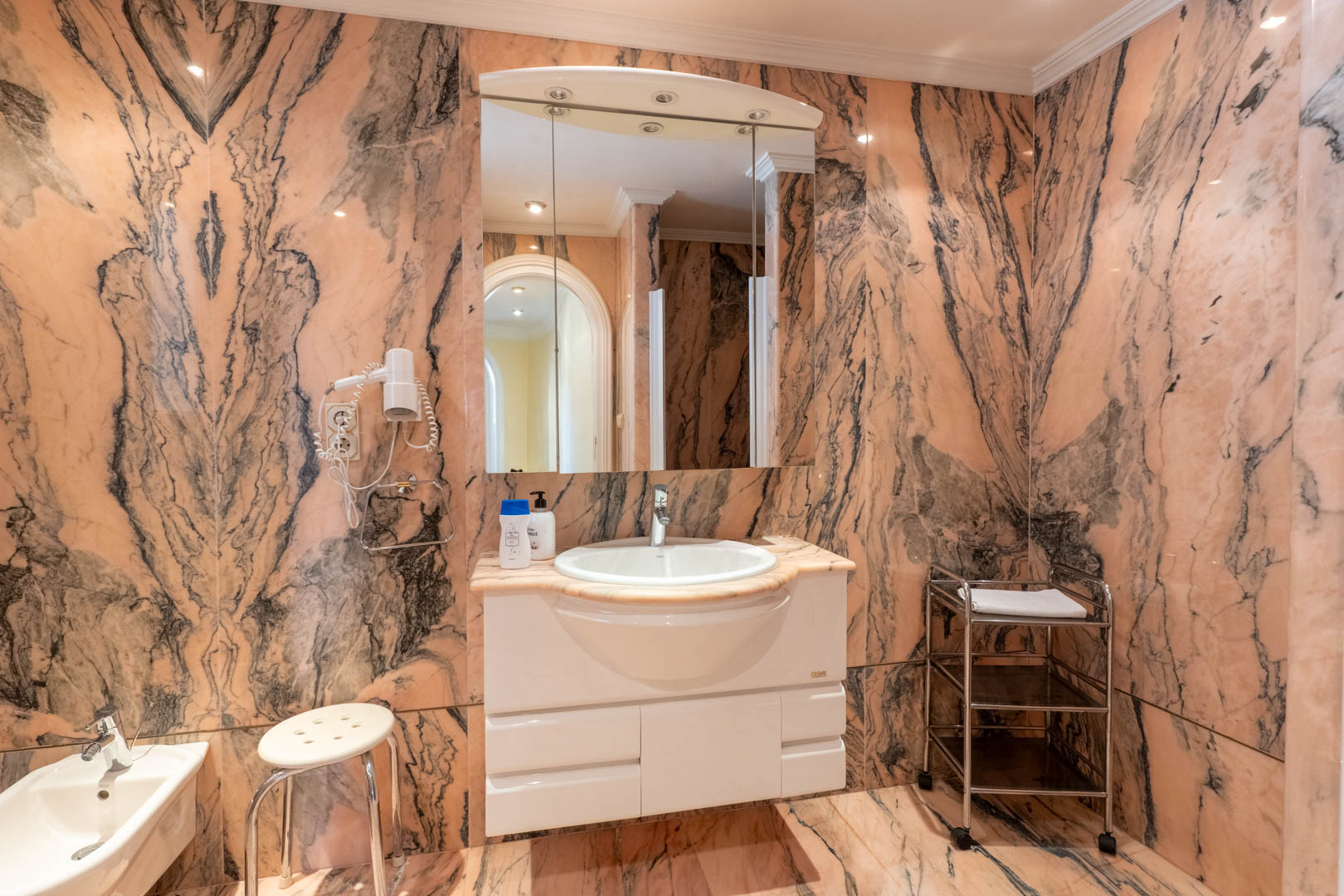 Badkamer met wanden en vloeren van roze marmer met donkere aders, voorzien van een wit wastafelmeubel, spiegelkast met verlichting en een bidet.