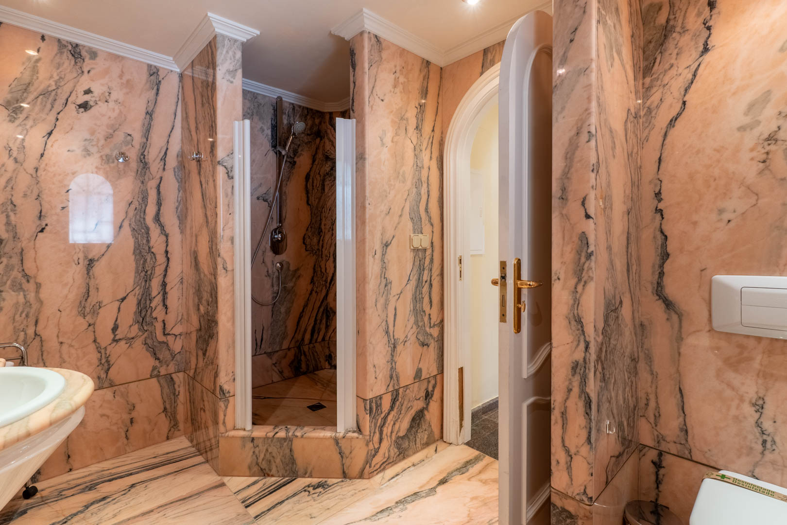 Badkamer volledig afgewerkt met roze geaderd marmer, voorzien van een inloopdouche, klassieke sierlijsten en een boogdeur met goudkleurig beslag.