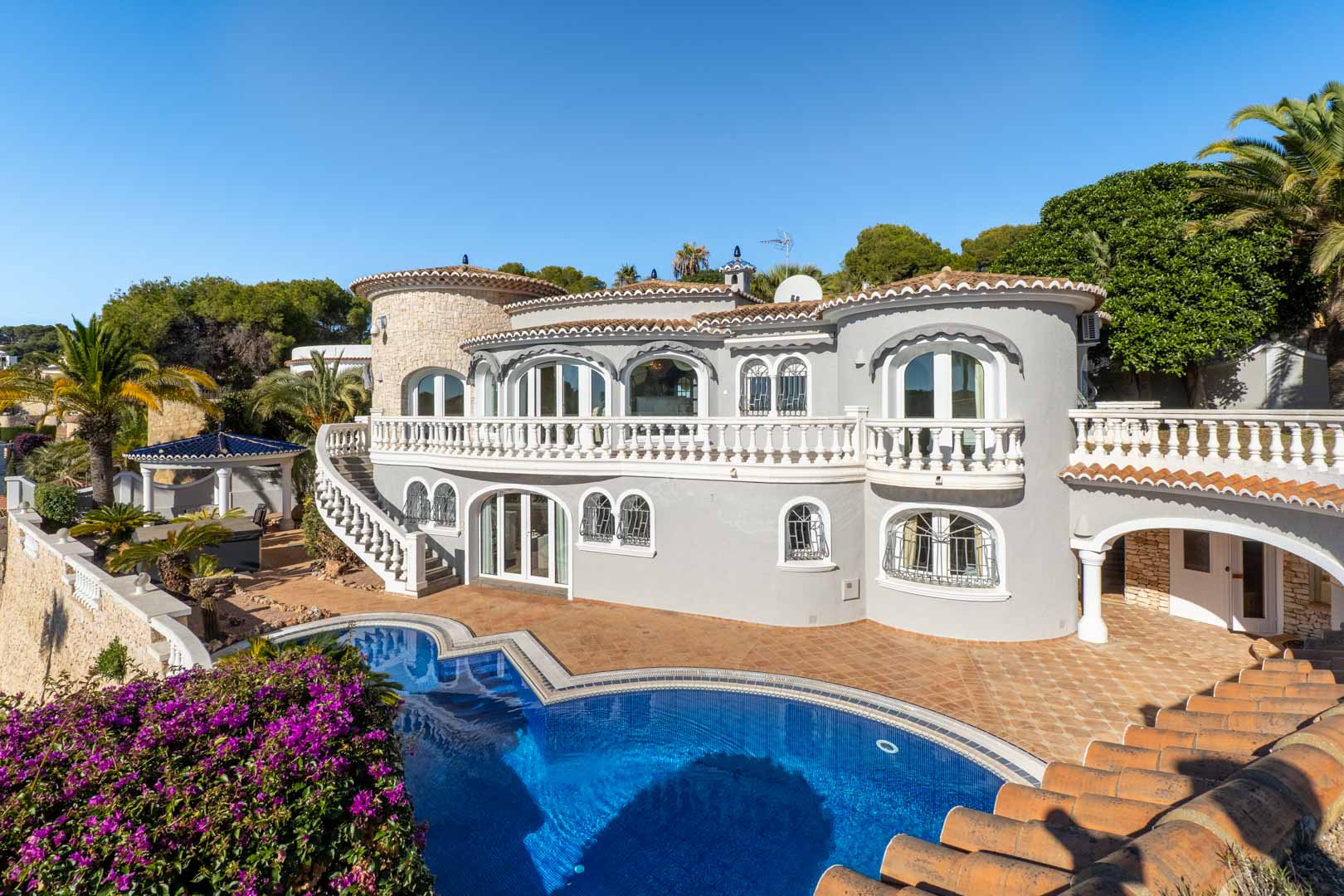 Buitenaanzicht van de villa met een gebogen zwembad, terrassen met witte balustrades en mediterrane architectuur in grijstinten onder een heldere hemel.