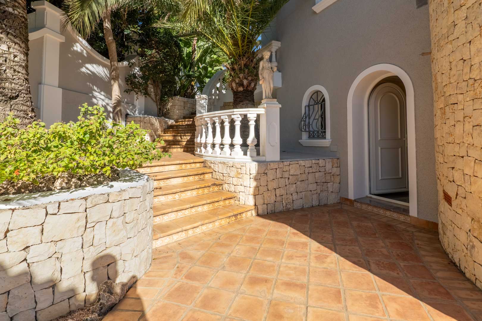 Buiteningang met terracotta vloer, natuurstenen muren en een boogdeur. Een stenen trap met balustrade leidt door de volgroeide mediterrane tuin met palmbomen.