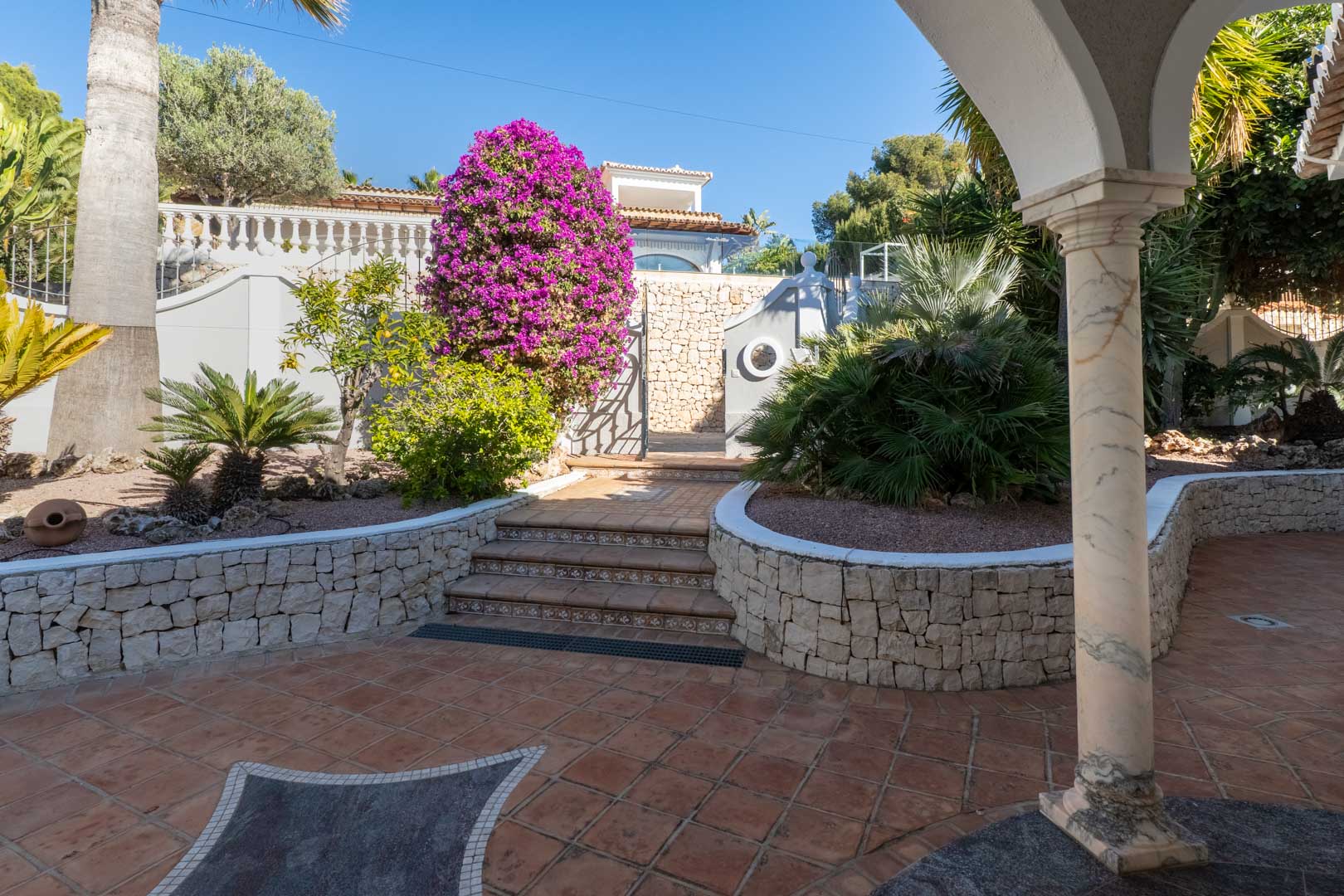 Buitenpatio met terracotta tegels, drooggestapelde muren en een bloeiende bougainville. Een marmeren zuil omlijst de trap naar de mediterrane tuinen.