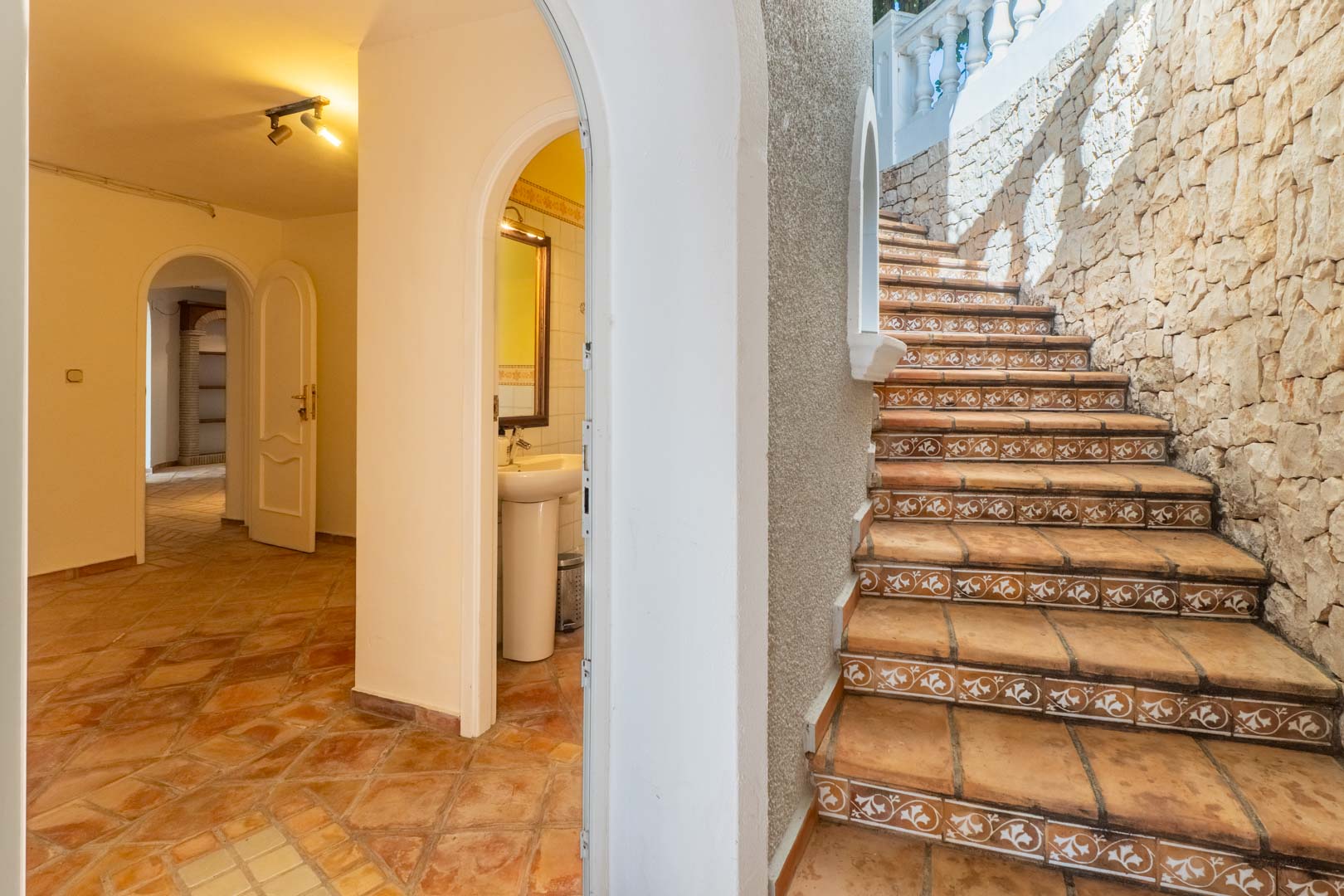 Buitentrap met decoratieve tegels en natuurstenen muur, grenzend aan een binnenhal met terracotta vloeren en gewelfde deuropeningen.