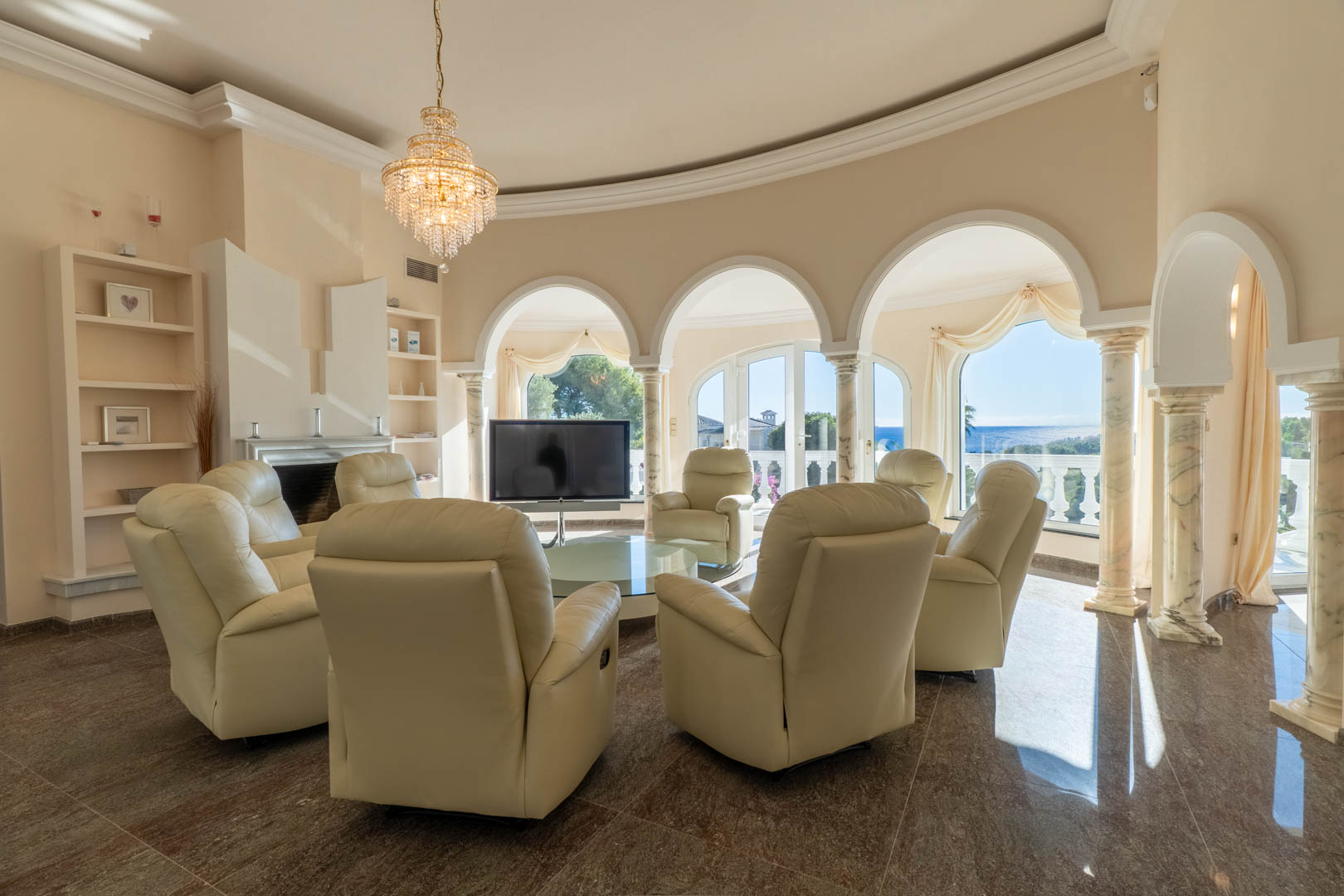 Circulaire woonkamer met boogvensters op marmeren zuilen, crème lederen fauteuils en panoramisch uitzicht op de Middellandse Zee.