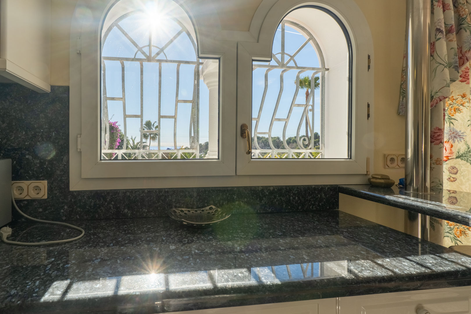 Donker granieten aanrechtblad onder boogvensters met decoratieve tralies, met uitzicht op de mediterrane tuin en palmbomen in het zonlicht.